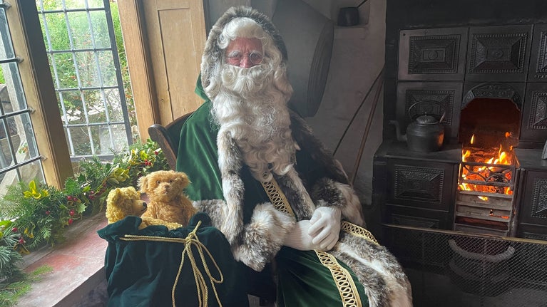 Father Christmas sits in a Rock House by a log fire, dressed in traditional green robes.